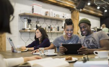 A group of people in an office breakroom. One reads and two look at a phone.