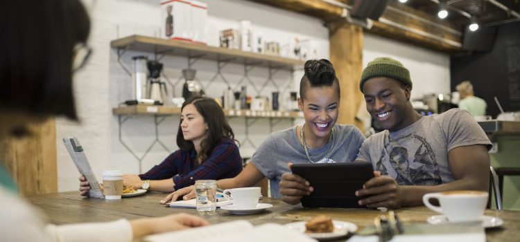 A group of people in an office breakroom. One reads and two look at a phone.