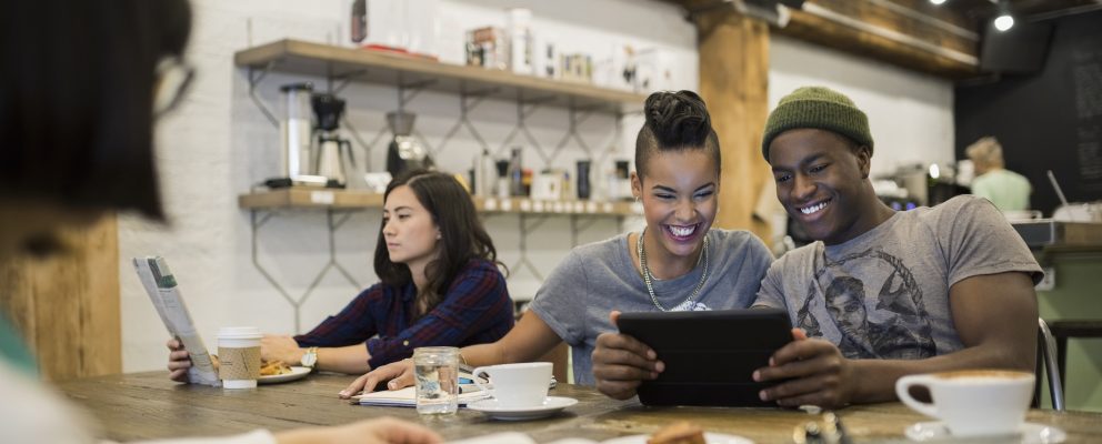 A group of people in an office breakroom. One reads and two look at a phone.
