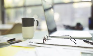 Financial papers, reading glasses, and laptop strewn about desk