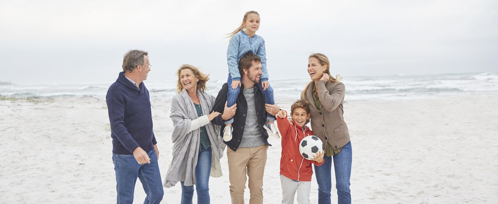 A family, including a grandmother, a grandfather, a mom, a dad, and two children, walk on a beach together.