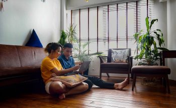 A man and a woman sit in the living room, reviewing their accounts on laptop computer.