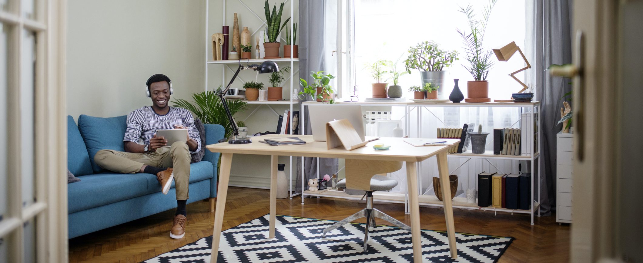 A man relaxes on a couch next to a home office desk, working on a tablet.