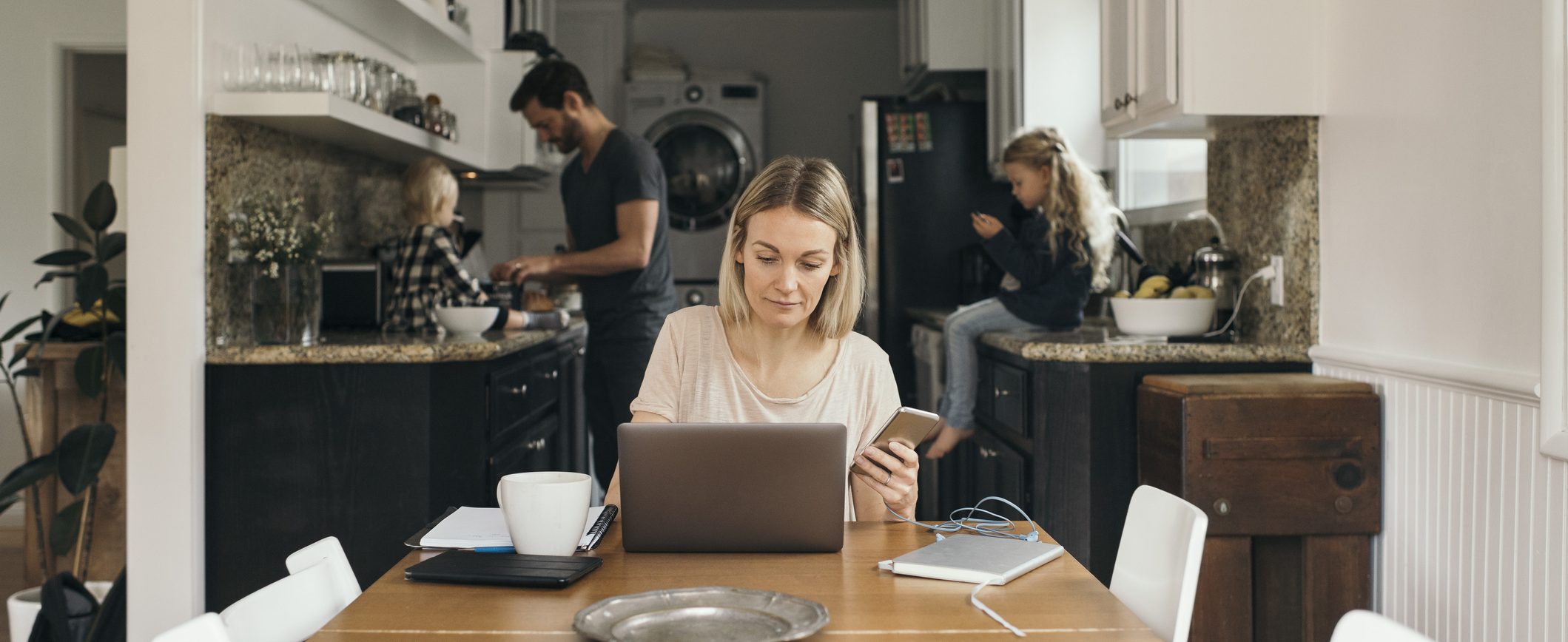 A woman works at the kitchen table on a laptop and smart phone while a man and two children occupy space in the back of the kitchen.