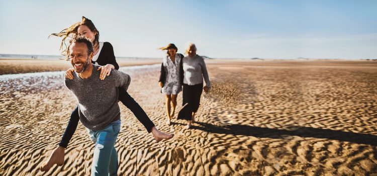 A man carries a woman on piggyback while walking on the beach, while two women walk behind with their arms around each other.