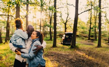 Man holds daughter and kisses his wife on the cheek in the woods.