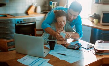 Couple looking at a mobile phone while reviewing paperwork in their kitchen