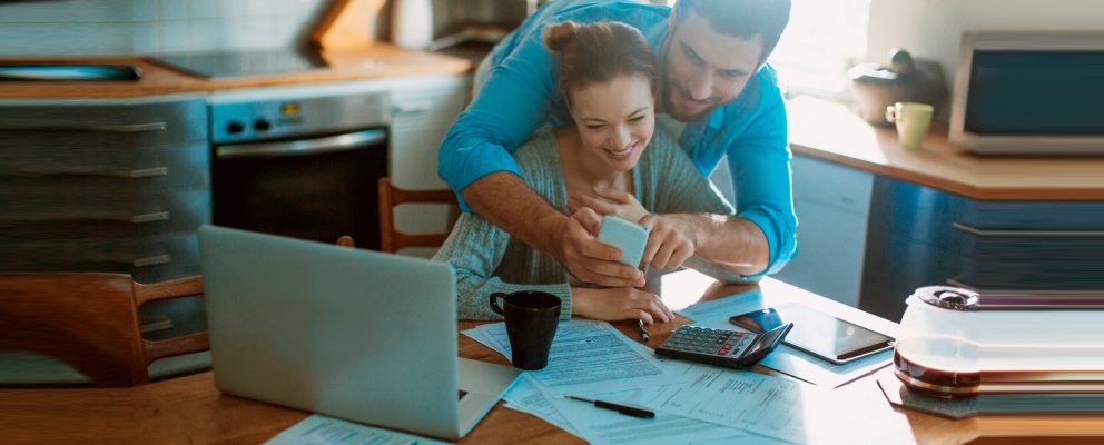 Couple looking at a mobile phone while reviewing paperwork in their kitchen