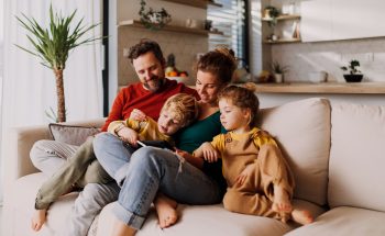 A family of four (mom, dad, a boy and a girl) sit on the couch and look at something.