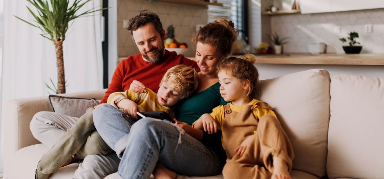 A family of four (mom, dad, a boy and a girl) sit on the couch and look at something.