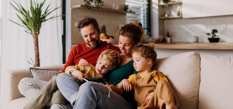 A family of four (mom, dad, a boy and a girl) sit on the couch and look at something.