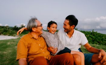 A grandpa, his son, and grandchild sit outside in the grass laughing.