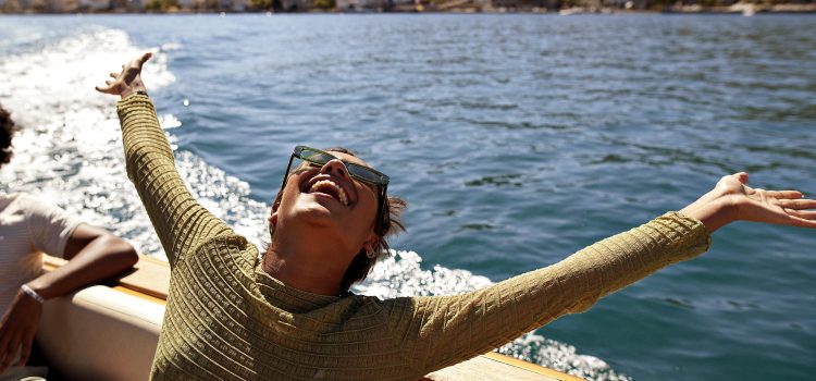 A woman stands by the ocean, tilting her head back and smiling up at the sky, her arms are open.