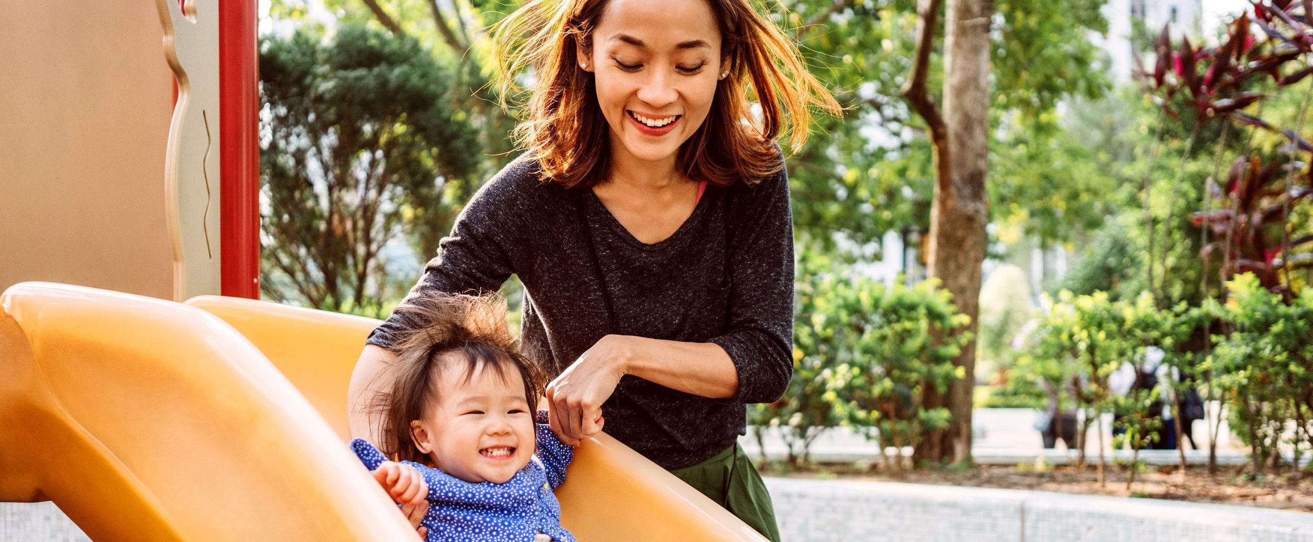 A woman holds hands with her small child as he slides down a slide.
