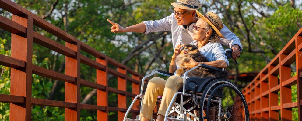On a bridge, a man stands next to a woman, who’s seated in a wheelchair with a dog in her lap.