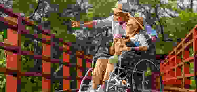 On a bridge, a man stands next to a woman, who’s seated in a wheelchair with a dog in her lap.
