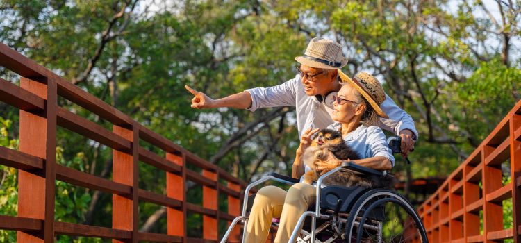 Older man points to something outside while on a walk with his wife who is in a wheelchair holding a dog.