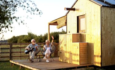 A retired couple sits on chairs outside. The man plays a guitar while the woman sings and drinks wine.