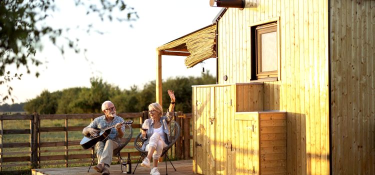 A retired couple sits on chairs outside. The man plays a guitar while the woman sings and drinks wine.