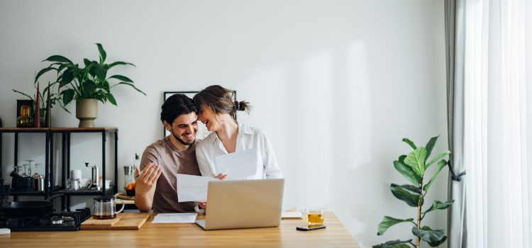 A couple stands close together while they review documents at the kitchen table.