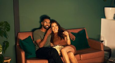 A couple sits on a couch while watching a movie and eating popcorn.