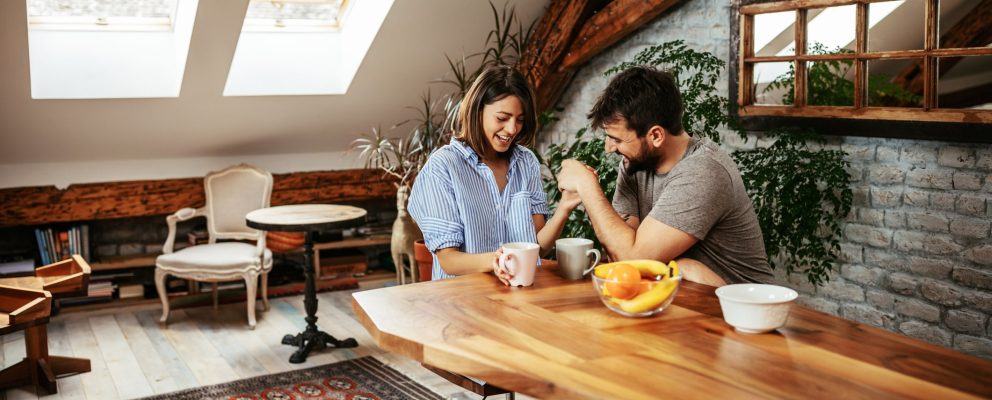 A man and woman laugh together while sitting at a dining table with cups of coffee.