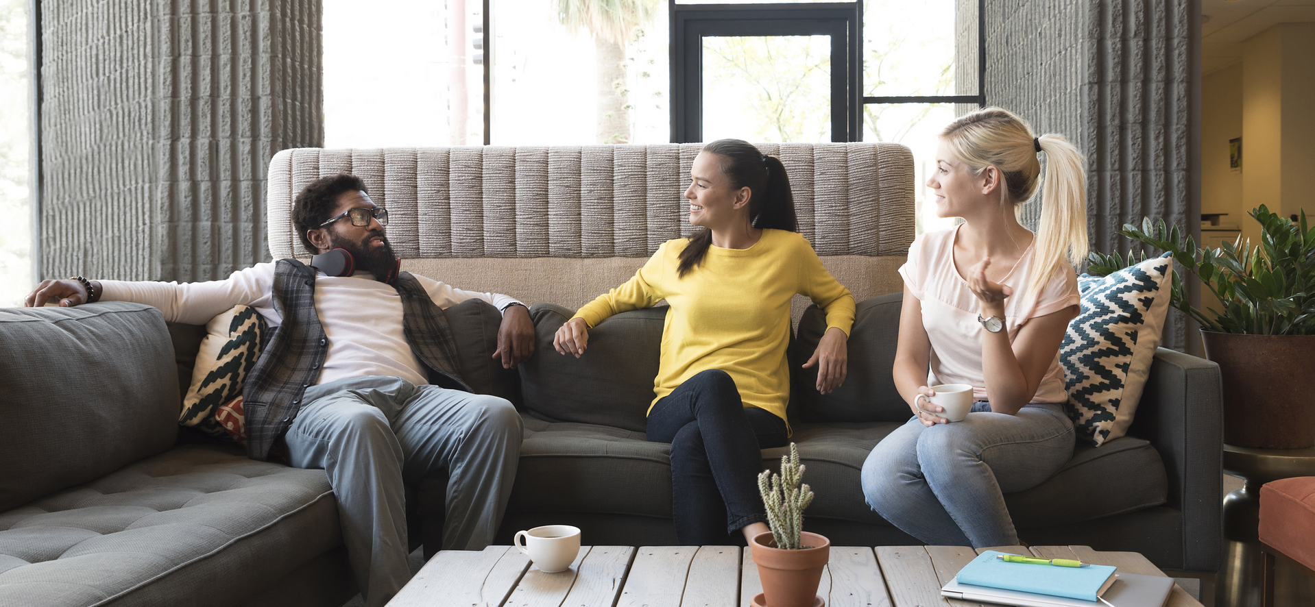 Three friends sit together on a sofa and chat, one holding a cup of coffee.