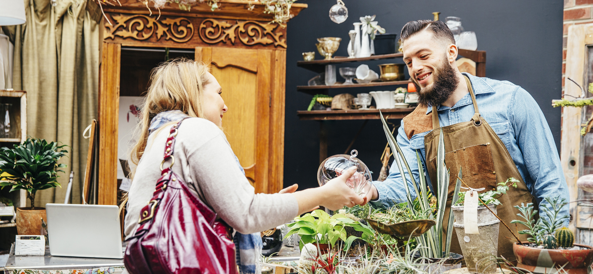In a plant shop, a woman examines a piece of glass with a man wearing an apron.