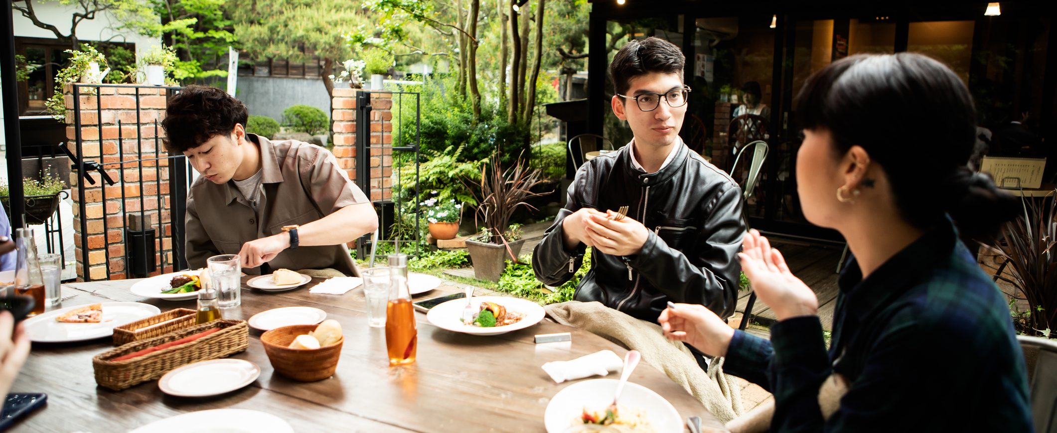 Three friends enjoy a meal at an outdoor table, with plants and trees in the background.