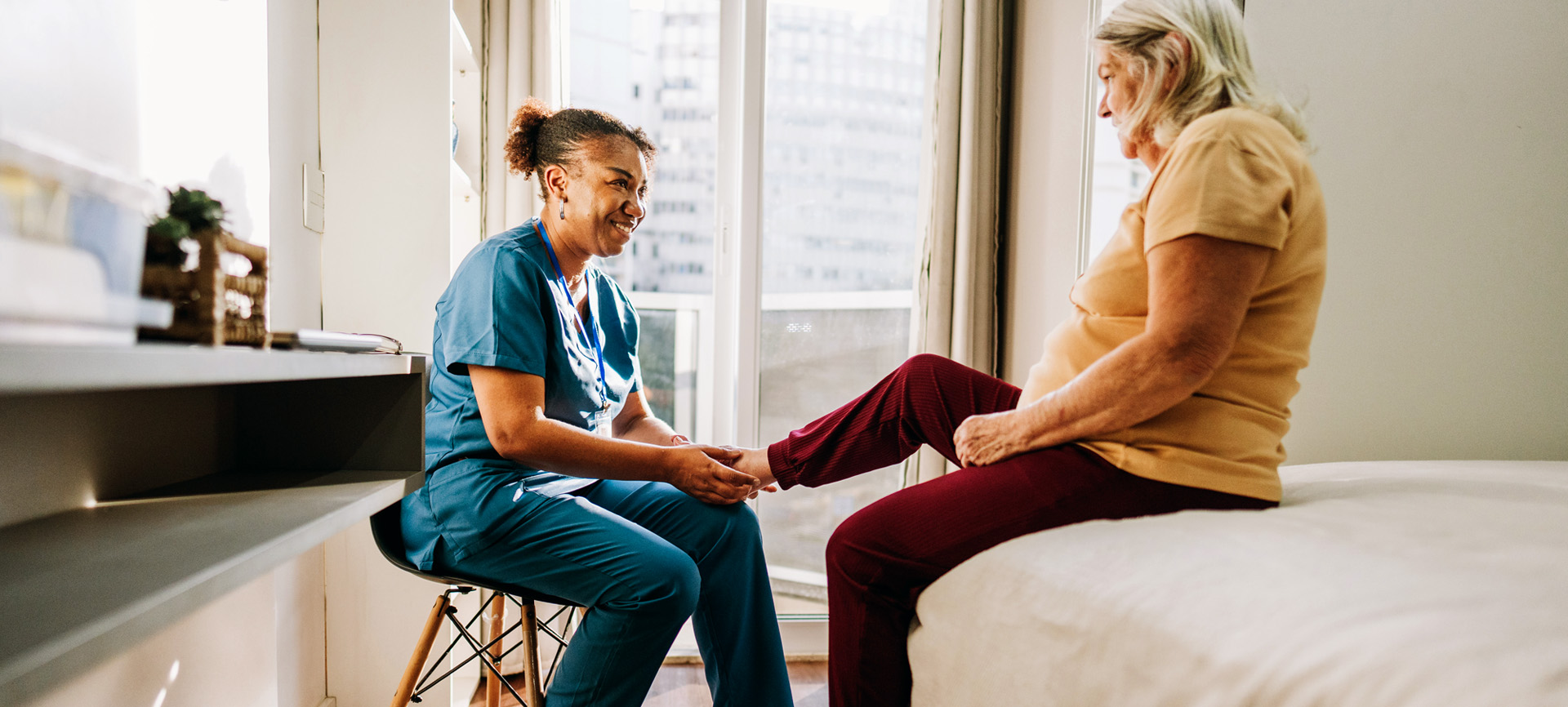 A nurse wearing blue scrubs assists an elderly woman sitting on a bed in a bright, sunny room. Both women smile.