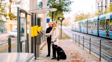 A blind woman uses a keypad outside of a train station while a man stands by her side.