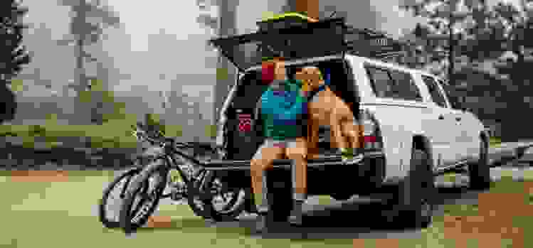 A man sits with his dog in an open car trunk, getting ready to hike a forest trail.