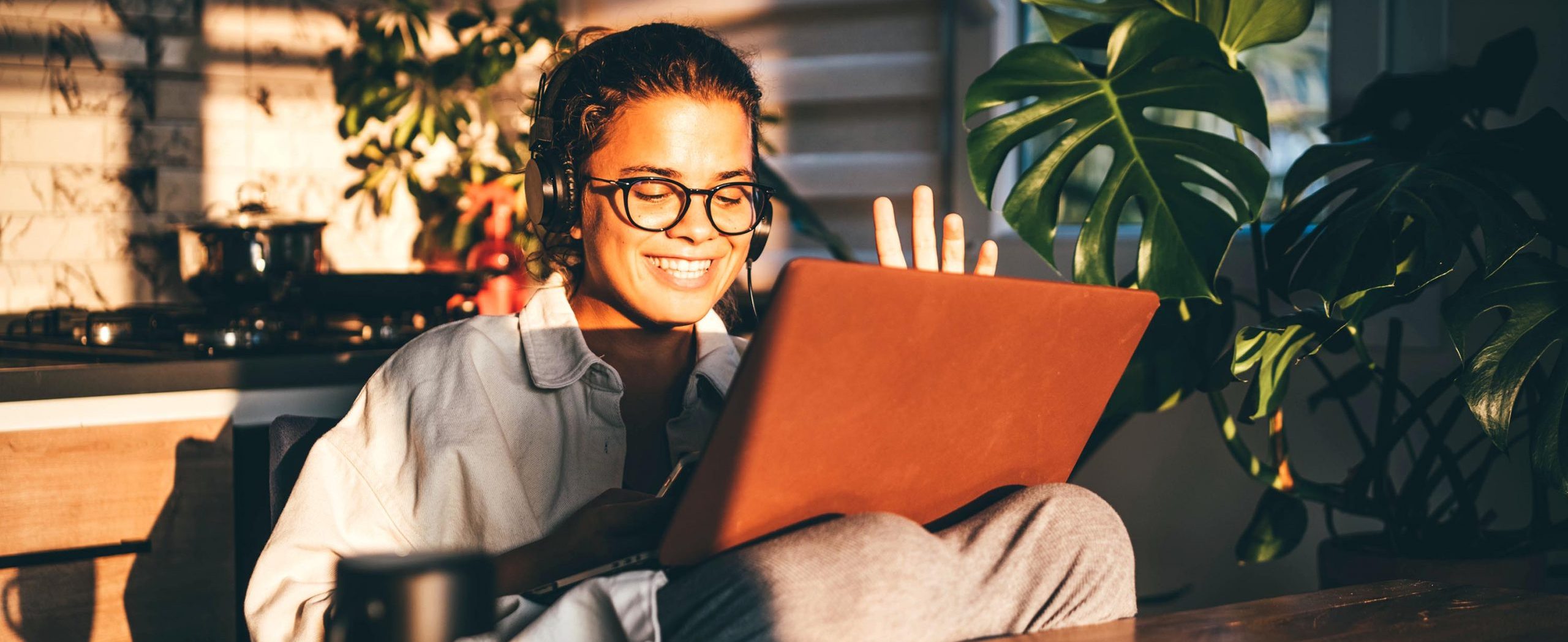 Sitting in her sunny kitchen with a cup of coffee, a woman smiles and waves at her laptop.