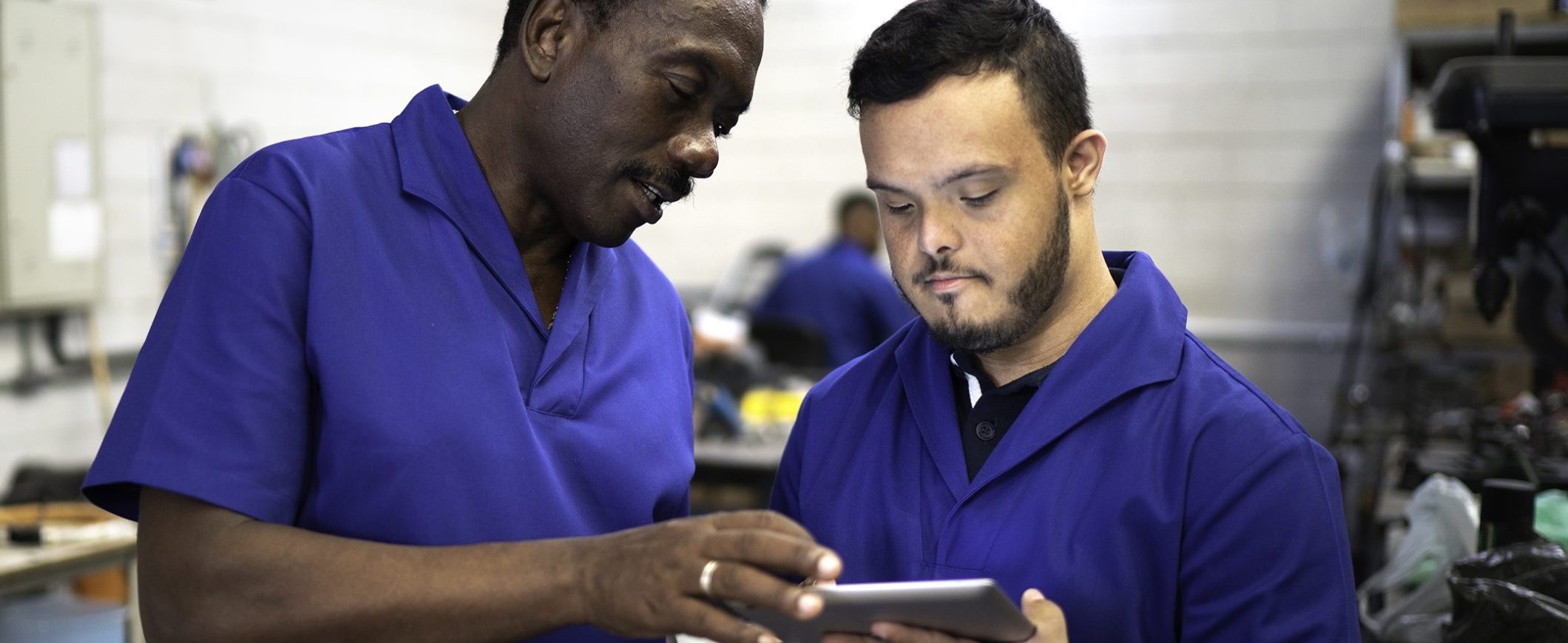 Two uniformed men in a factory stand together and look at a tablet.