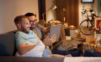 A couple sits together on a couch and looks at a tablet, with a bike and potted plants in the background.