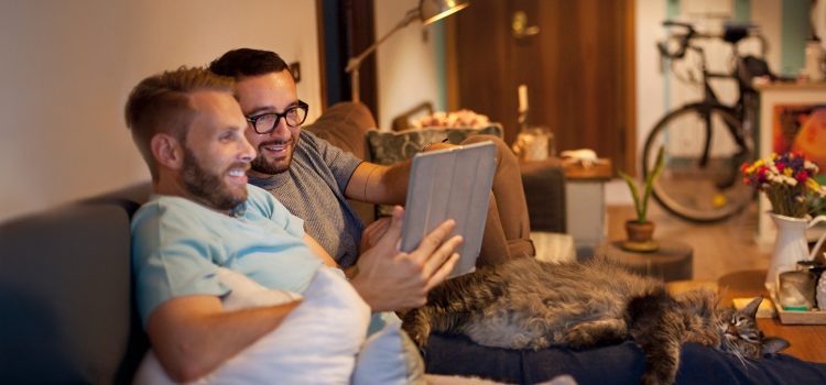A couple sits together on a couch and looks at a tablet, with a bike and potted plants in the background.