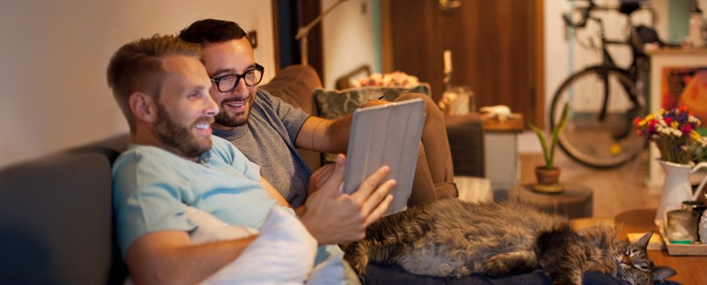 A couple sits together on a couch and looks at a tablet, with a bike and potted plants in the background.