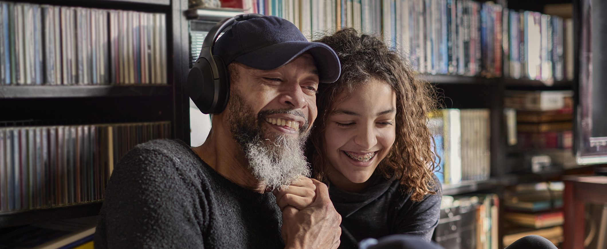 A man and his young daughter share headphones and listen to music together in front of shelves of CDs.