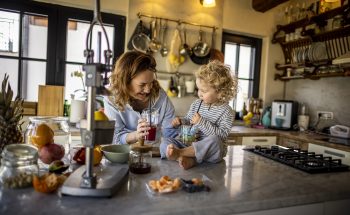 A woman stands in her kitchen holding a drink with straw, next to her young daughter who sits on the counter.