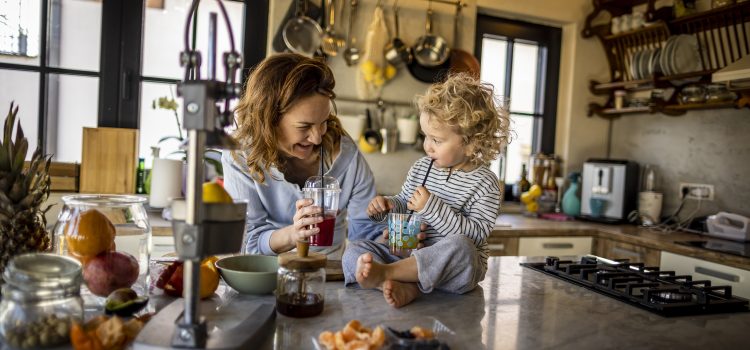 A woman stands in her kitchen holding a drink with straw, next to her young daughter who sits on the counter.