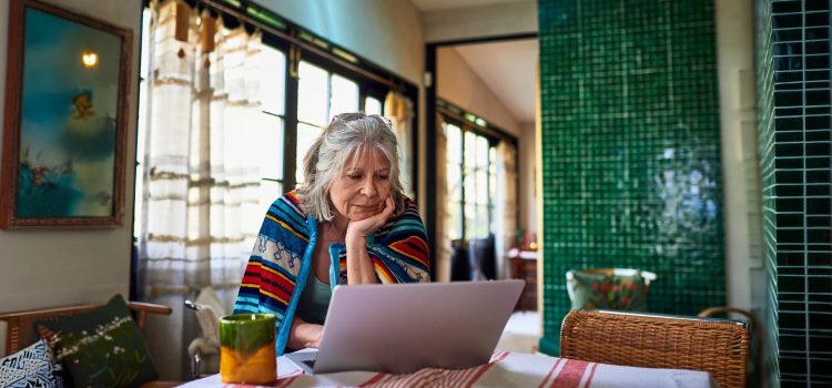 A woman sits at her dining table, resting her chin in her hand as she looks at her laptop.