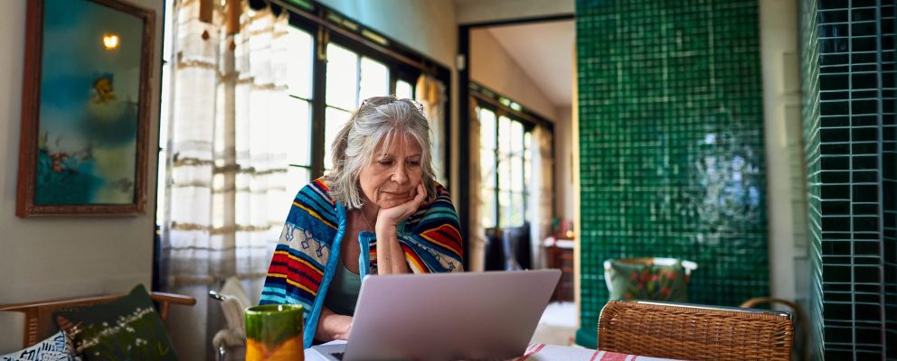A woman sits at her dining table, resting her chin in her hand as she looks at her laptop.