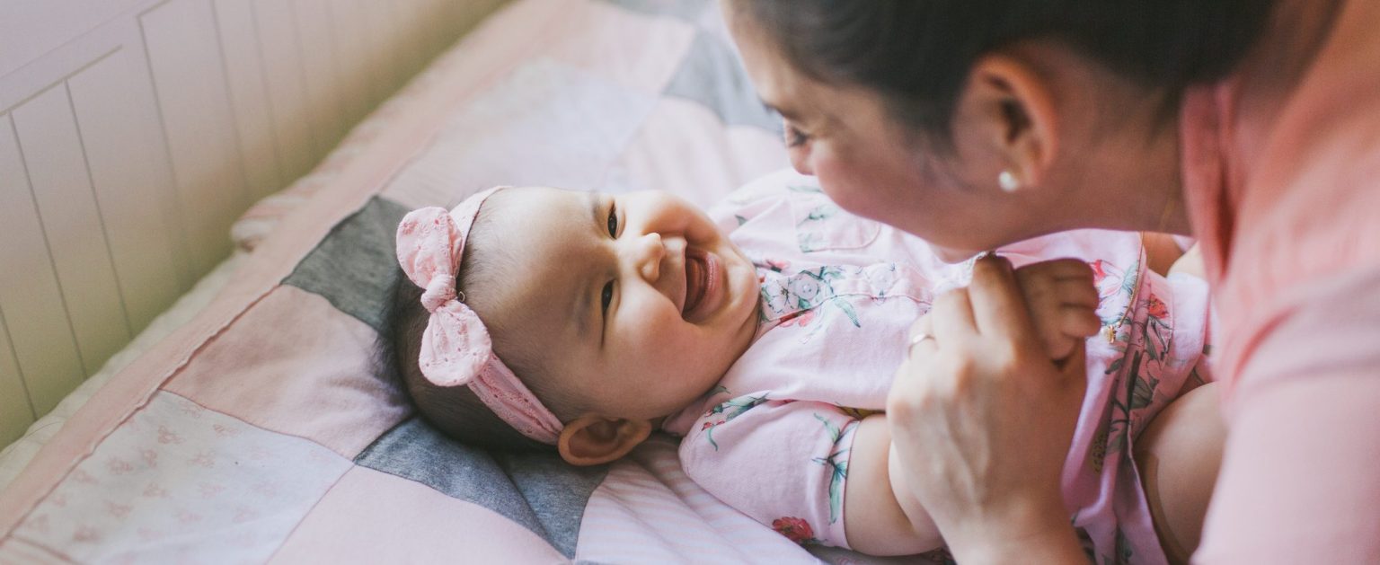 A baby girl on a pink and gray quilt grins at her mom who’s also smiling and holding one of the baby’s hands.