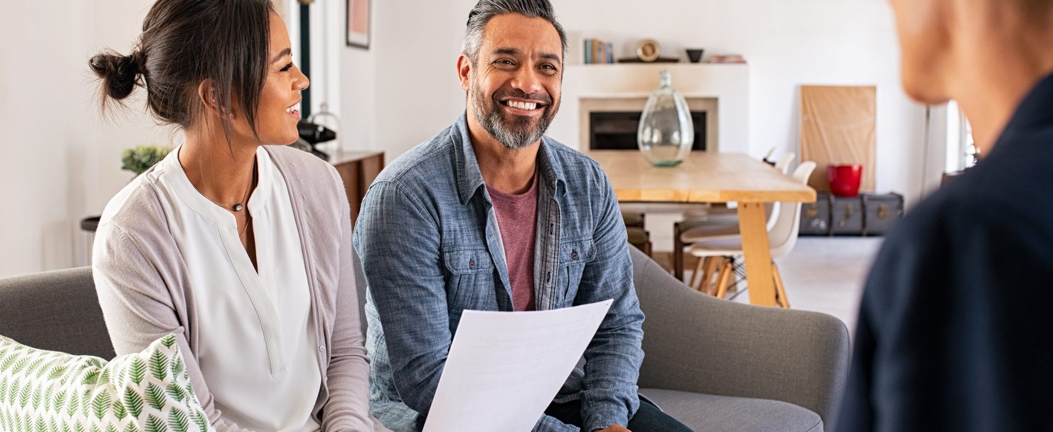 A couple sits on the couch in their living room with a guest, looking over paperwork together.