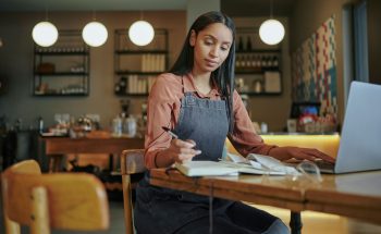 A young woman in a denim apron sits in a workspace, focused on paperwork while using a laptop.