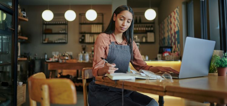A young woman in a denim apron sits in a workspace, focused on paperwork while using a laptop.