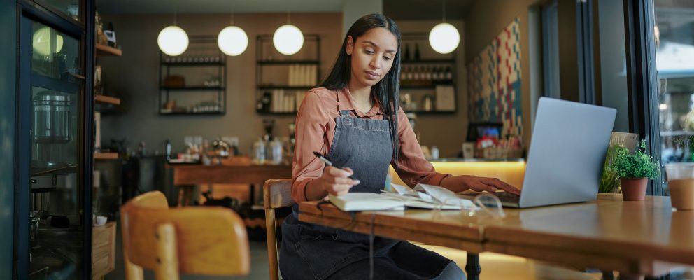 A young woman in a denim apron sits in a workspace, focused on paperwork while using a laptop.