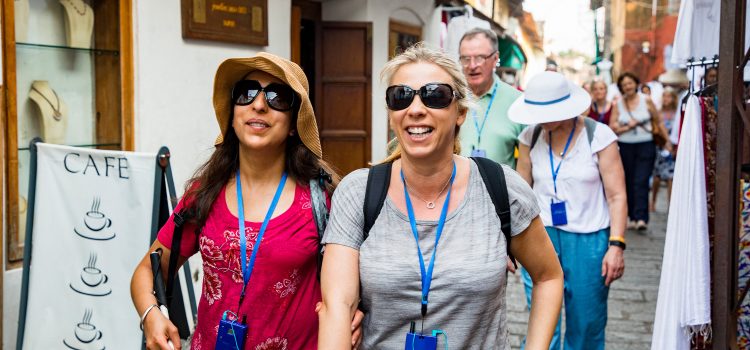 A woman wearing sunglasses and holding a cane walks arm-in-arm with a friend on a city street, accompanied by their tourist group.