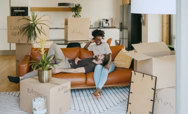 A couple relaxes on a couch surrounded by unpacked moving boxes in their living room.