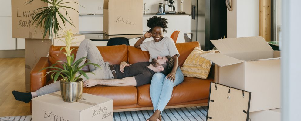 A couple relaxes on a couch surrounded by unpacked moving boxes in their living room.